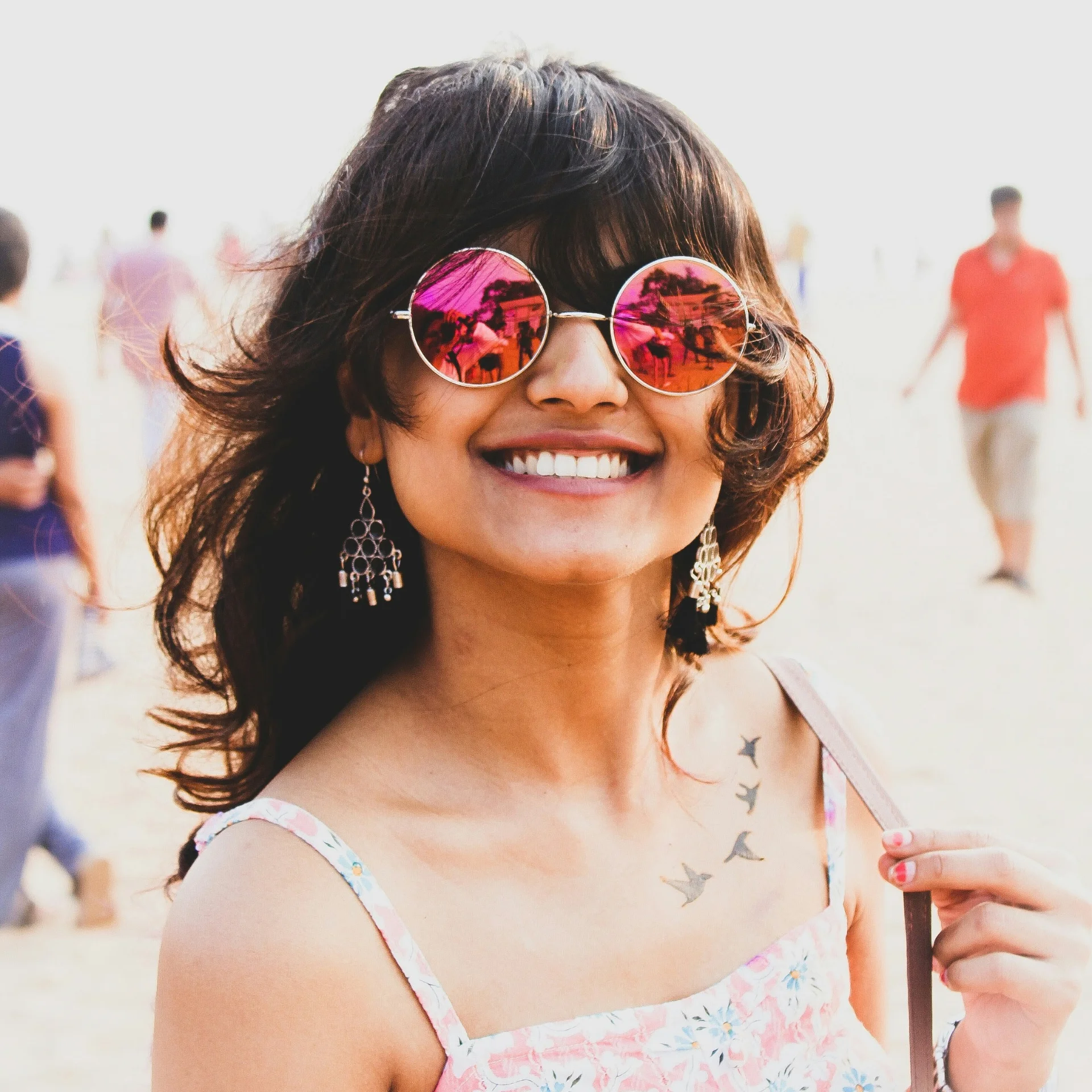 A woman wearing sunglasses and dangly earrings smiles in a beachy location